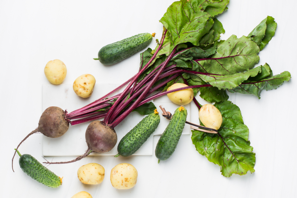 Frisches Wurzelgemüse mit Kartoffeln, Gurken und Rote-Bete-Blättern auf weißem Untergrund – intuitives Kochen mit saisonalen Zutaten.